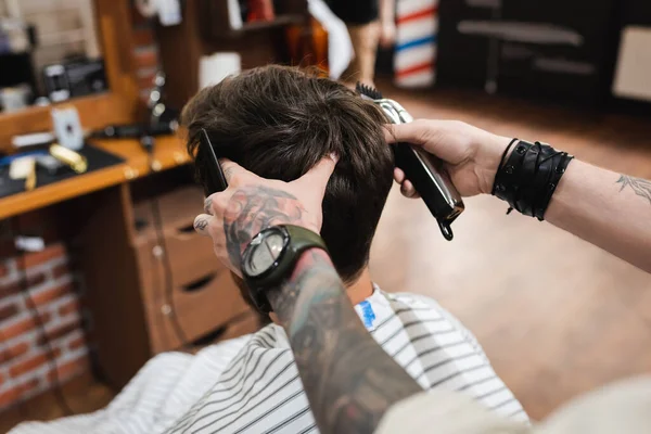 Tatuato barbiere in orologio da polso tenendo i capelli clipper e pettine vicino bruna uomo — Foto stock