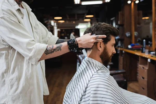 Vista lateral do barbeiro tatuado perto de cliente morena na capa de cabeleireiro — Fotografia de Stock