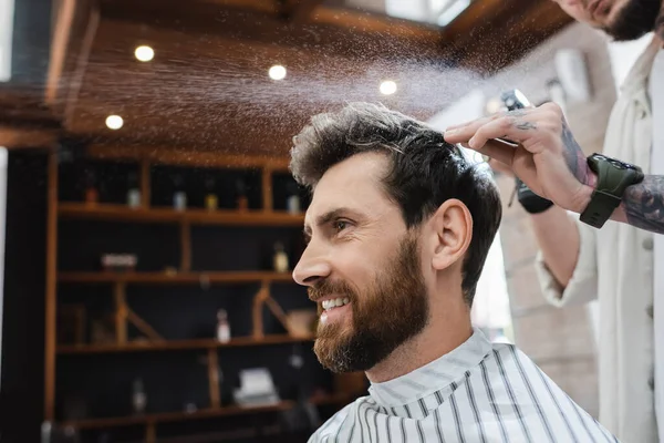 Barbudo morena hombre sonriendo cerca peluquería aplicando spray para el cabello - foto de stock