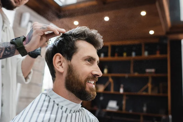Vista lateral del hombre barbudo alegre cerca de peluquero aplicando spray para el cabello - foto de stock