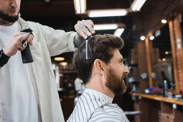 Татуированный парикмахер держит бутылку лака для волос и расчесывает клиента брюнетки — стоковое фото