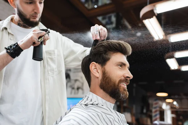 Felice uomo bruna vicino barbiere applicare lacca per capelli nel salone di bellezza — Foto stock