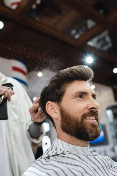 Брюнетка людина з бородою посміхається біля перукаря, застосовуючи спрей для волосся — стокове фото