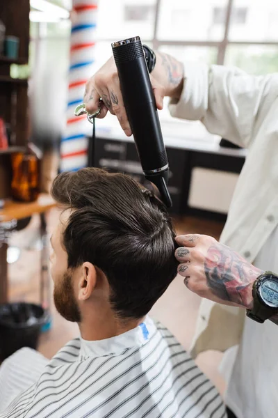 Coiffeur tatoué dans la montre-bracelet séchage des cheveux du client dans le salon de coiffure — Photo de stock