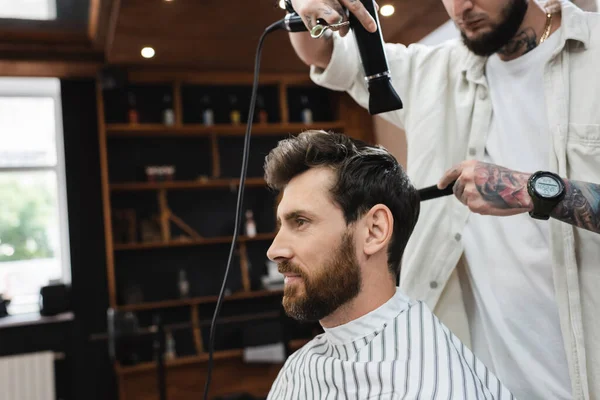 Bearded man in hairdressing cape near barber drying his hair in beauty salon — Stock Photo