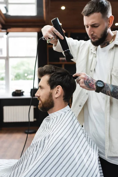 Tattooed hairdresser drying hair of brunette man in barbershop — Stock Photo