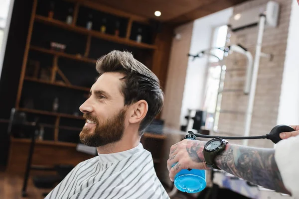 Vista lateral del hombre barbudo feliz en la capa de peluquería cerca de barbería rociar perfume - foto de stock