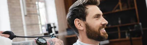 Tatuado cabeleireiro pulverização perfume no homem barbudo satisfeito na barbearia, banner — Fotografia de Stock
