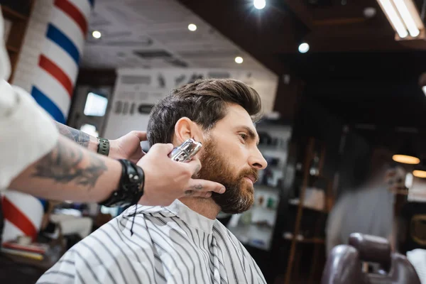 Peluquería borrosa sosteniendo afeitadora eléctrica cerca del hombre barbudo en la barbería - foto de stock