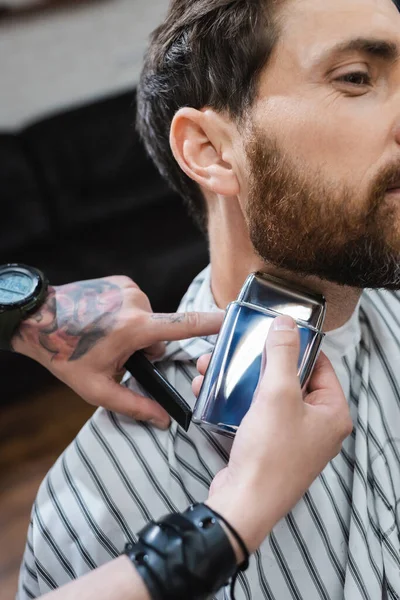 Vista cortada de barbeiro barbeando pescoço de homem barbudo com navalha elétrica — Fotografia de Stock