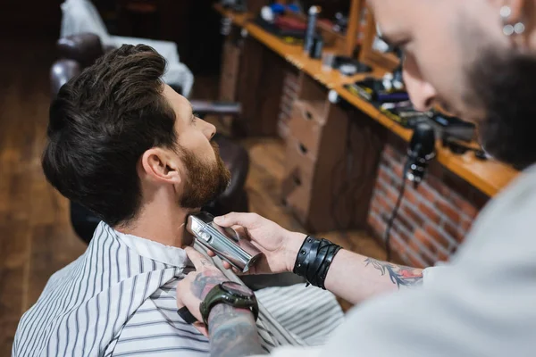 Tattooed barber shaving neck of brunette man with electric razor on blurred foreground — Stock Photo