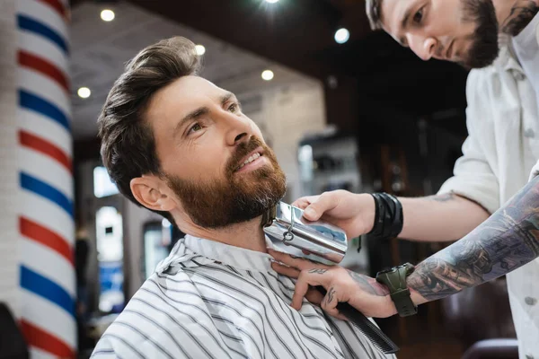Бородатый мужчина улыбается, когда парикмахер бреет шею электрической бритвой — стоковое фото