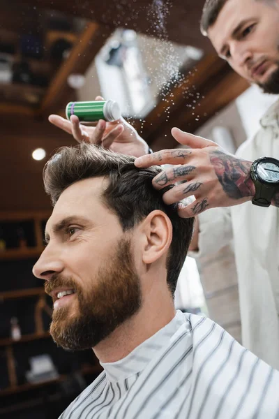 Hombre barbudo sonriendo cerca de peluquero borroso aplicando polvo de textura en su cabello - foto de stock