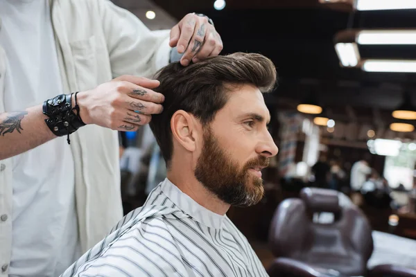 Morena barbudo homem no cape cabeleireiro perto tatuado barbeiro tocando seu cabelo no salão de beleza — Fotografia de Stock