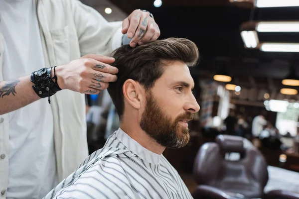 Brunette bearded man in hairdressing cape near tattooed barber touching his hair in beauty salon — Stock Photo