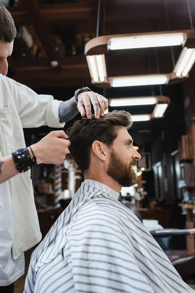 Vista laterale di uomo bruna barbuto in mantello parrucchiere vicino barbiere in salone di bellezza — Foto stock
