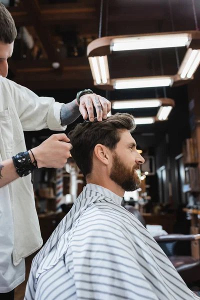 Vista lateral de hombre morena barbuda en capa de peluquería cerca de peluquero en salón de belleza - foto de stock