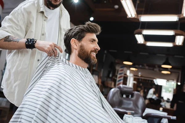 Peluquero poniendo capa de peluquería en cliente feliz en la barbería - foto de stock