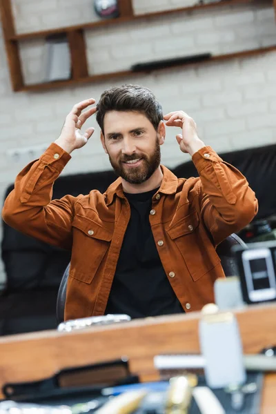Pleased bearded man looking in mirror and touching hair in beauty salon — Stock Photo