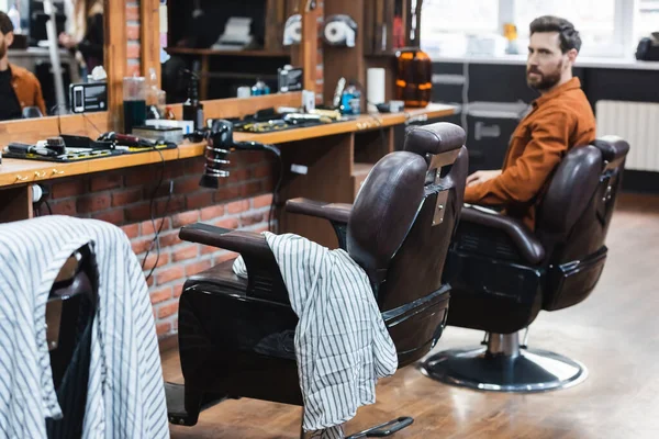 Hombre barbudo sentado en silla de peluquería y mirando a la cámara en la barbería - foto de stock