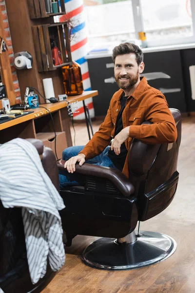Homem barbudo feliz sentado na barbearia e sorrindo para a câmera — Fotografia de Stock