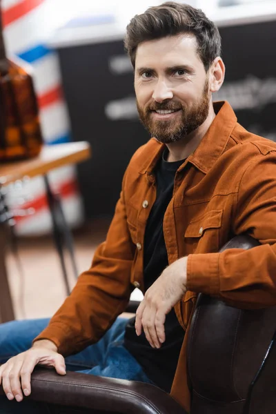 Hombre barbudo sonriendo a la cámara mientras está sentado en la peluquería borrosa - foto de stock