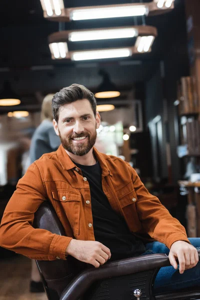 Homem barbudo alegre olhando para a câmera enquanto sentado na barbearia — Fotografia de Stock