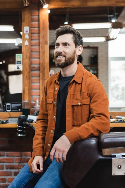 Улыбающийся бородатый мужчина сидит на парикмахерском кресле возле размытых зеркал в парикмахерской — стоковое фото