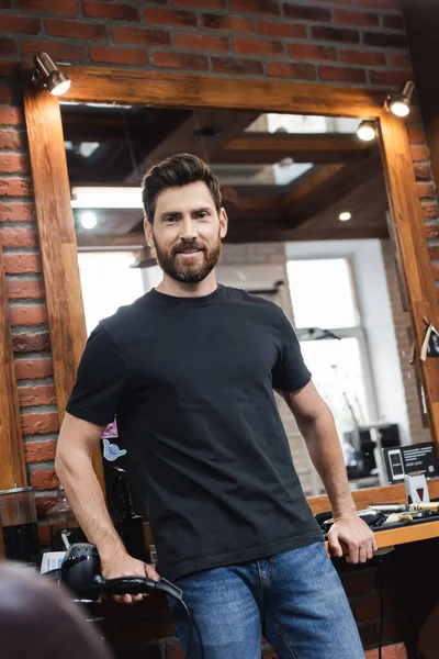 Cabeleireiro barbudo positivo sorrindo para a câmera perto do espelho na barbearia — Fotografia de Stock