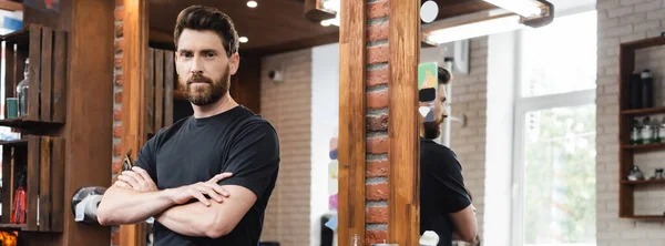 Cabeleireiro morena de pé com braços cruzados perto de espelhos na barbearia, bandeira — Fotografia de Stock