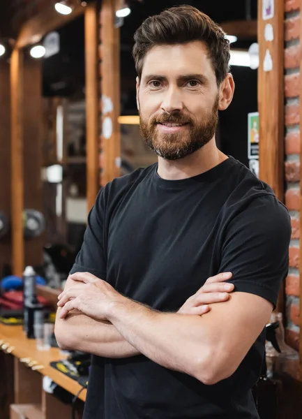 Morena peluquero barbudo de pie con los brazos cruzados en la barbería - foto de stock