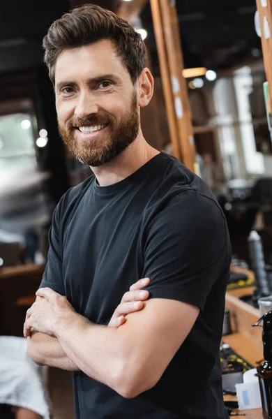 Joyful brunette barber standing with crossed arms and looking at camera in beauty salon — Stock Photo