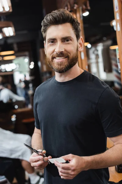 Heureux barbier tenant ciseaux et peigne tout en regardant la caméra dans le salon de beauté — Photo de stock