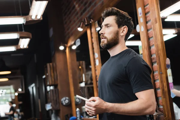 Brunette bearded hairstylist with scissors and comb near blurred mirrors in barbershop — Stock Photo