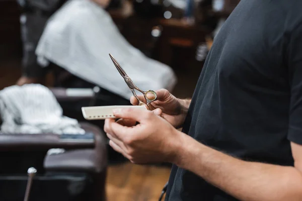 Cropped view of hairdresser holding comb and scissors in blurred barbershop — Stock Photo