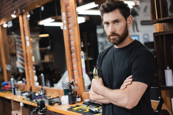 Barbe barbier debout avec les bras croisés et tondeuse à cheveux près de miroirs flous dans le salon de beauté — Photo de stock