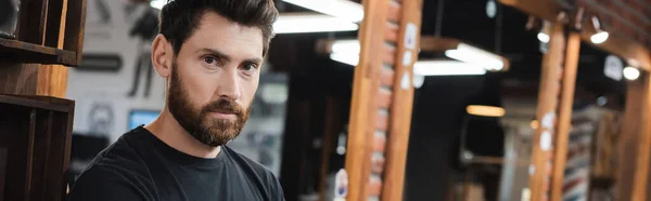 Bearded hairdresser looking at camera near blurred mirrors in barbershop, banner — Stock Photo