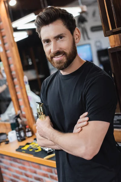 Glücklicher Friseur mit Haarschneidemaschine, während er im Friseurladen in die Kamera lächelt — Stockfoto