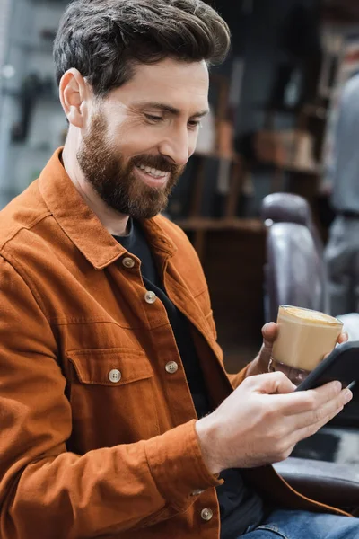 Homme barbu gai avec verre de cappuccino bavarder sur smartphone dans le salon de coiffure — Photo de stock