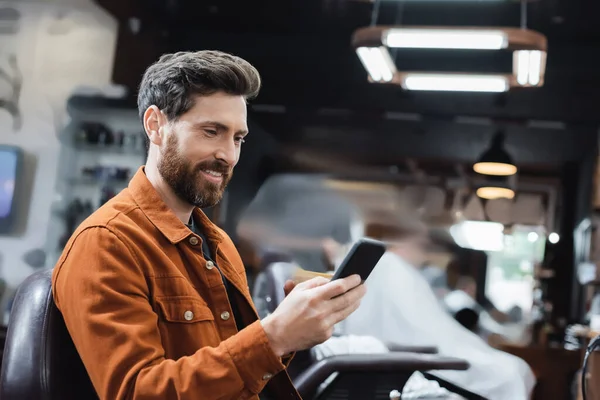 Homem barbudo feliz usando telefone celular na barbearia borrada — Fotografia de Stock