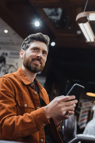 Homem barbudo sorrindo usando telefone celular na barbearia e olhando para longe — Fotografia de Stock