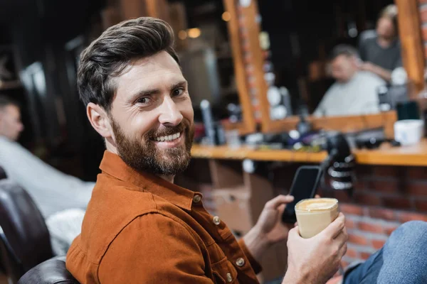 Homme barbu avec verre de cappuccino et téléphone cellulaire flou souriant à la caméra dans le salon de coiffure — Photo de stock