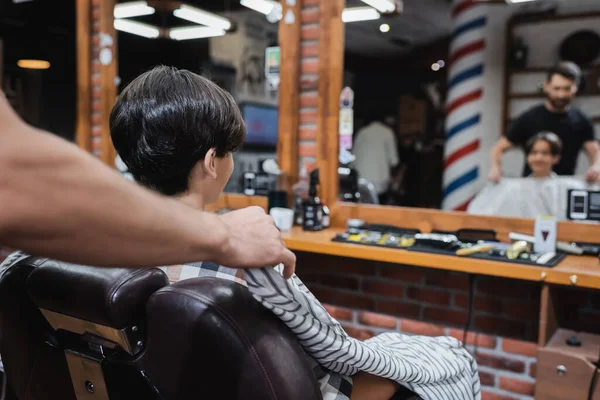 Coiffeur mettre coiffure cape sur adolescent client assis dans fauteuil près de miroirs flous — Photo de stock