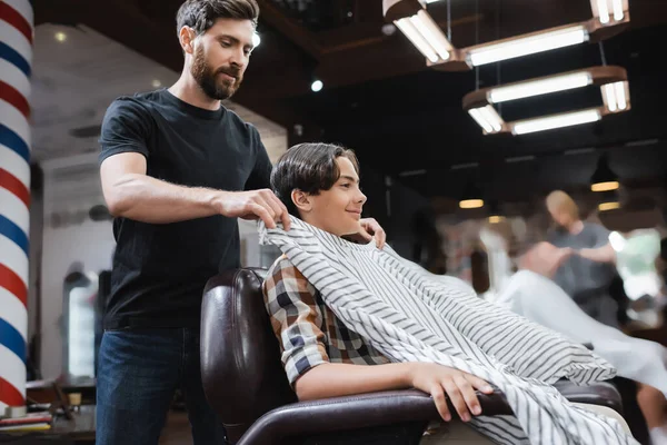 Бородатий перукар кладе перукарську накидку на підліткового клієнта, посміхаючись у кріслі — стокове фото