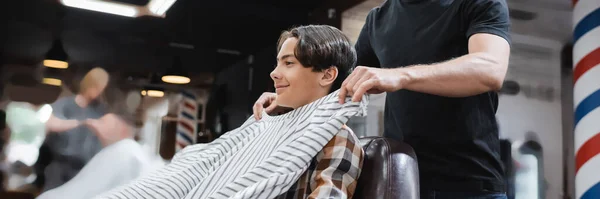 Vista lateral do barbeiro colocando capa de cabeleireiro no menino adolescente sorridente no salão de beleza, banner — Fotografia de Stock