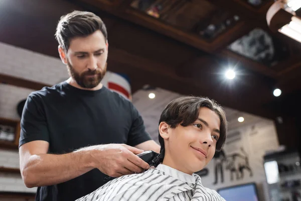 Adolescente chico en peluquería capa sonriendo mientras peluquero recortar su cabello en salón de belleza - foto de stock