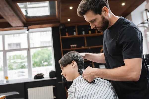 Вид збоку бородатий перукар обрізає волосся підлітка-хлопчика з електричним затискачем для волосся — стокове фото
