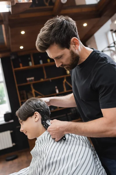 Vista lateral del peluquero barbudo recortando el cabello de un adolescente en la capa de peluquería - foto de stock