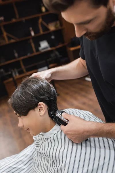 Cabeleireiro desfocado aparando o cabelo do cliente adolescente com cortador de cabelo — Fotografia de Stock