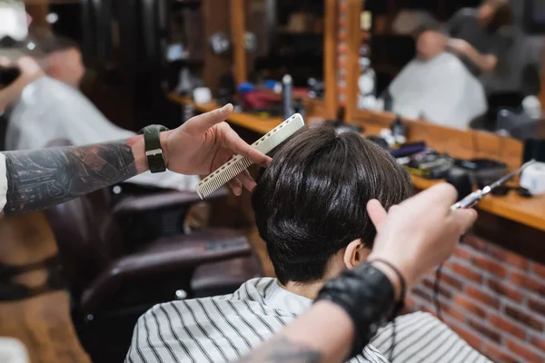 Peluquero tatuado con peine y tijeras borrosas cerca de adolescente morena en salón de belleza - foto de stock